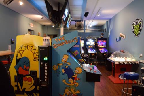 A retro arcade featuring "Pac-Man" and "The Simpsons" machines, with colorful decor and various games in the background.