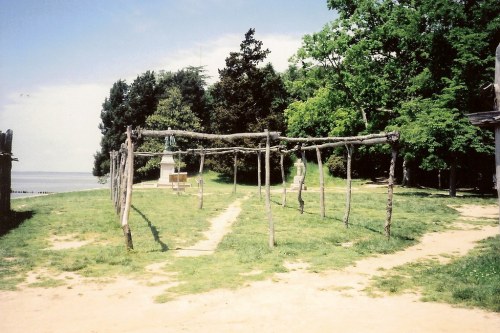 A grassy area with wooden posts forming a structure, surrounded by trees and a path leading to a water view.