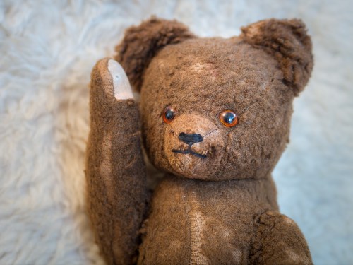 A worn, brown teddy bear with button eyes and a raised paw, resting on a soft, textured surface.