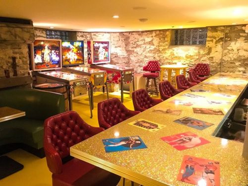 A retro game room featuring pinball machines, a glittery bar, and red tufted seating against a stone wall.