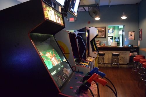 A row of vintage arcade machines in a retro gaming bar, with colorful screens and controllers visible.