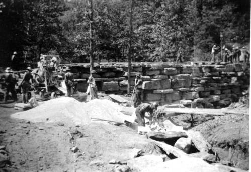 Workers constructing a stone wall in a forested area, with equipment and materials scattered around.