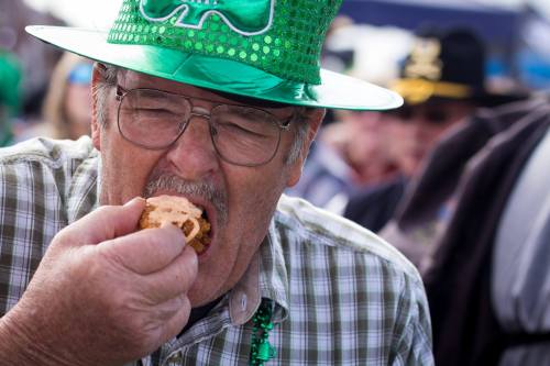 A man in a green hat bites into a sandwich, celebrating at a festive outdoor event.