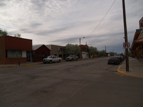 A quiet street scene with parked cars and small buildings under a cloudy sky.