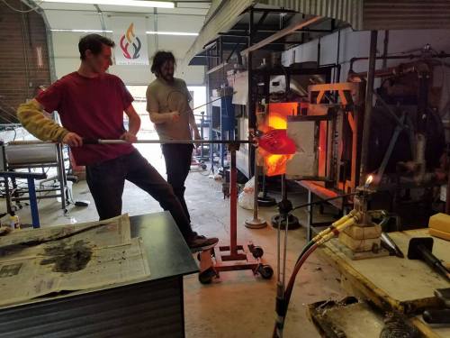 Two artists working in a glassblowing studio, one shaping molten glass while the other assists in the background.