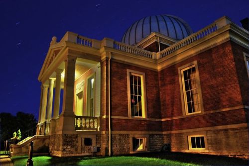 A historic brick building with a dome, illuminated at night, surrounded by green grass and starry sky.