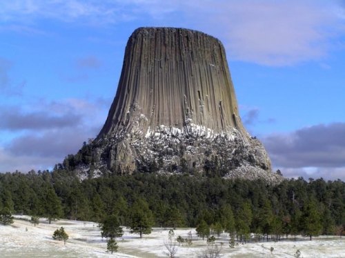 A towering rock formation with vertical stripes, surrounded by trees and a snowy landscape under a blue sky.