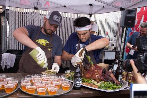 Two chefs prepare a whole roasted pig on a table, surrounded by drinks and small cups, under a tent.