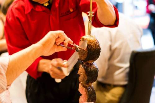 A server in a red shirt slices meat from a skewer at a restaurant, while a diner holds a plate ready to serve.
