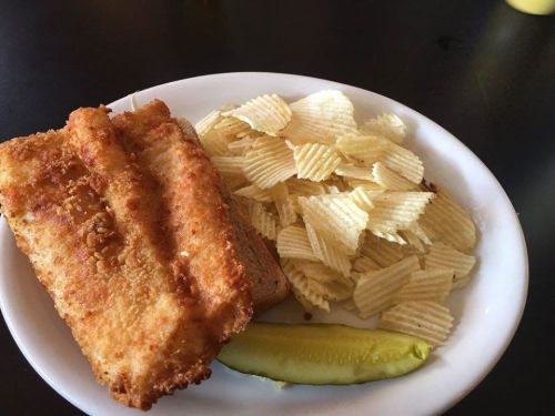 A plate with a fried fish sandwich, potato chips, and a pickle slice on the side.