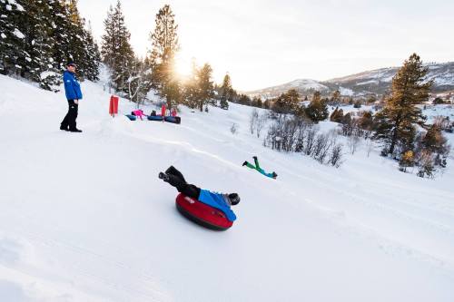 A snowy landscape with people tubing down a hill, surrounded by trees and mountains, under a bright sunset.