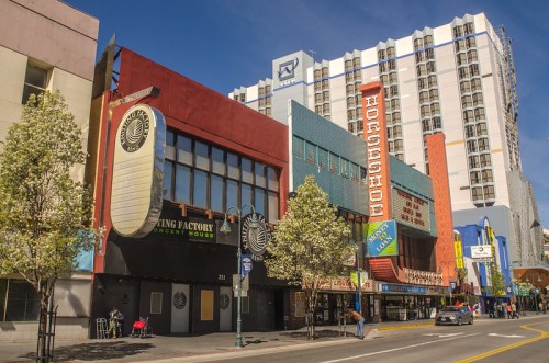 Colorful buildings line a city street, featuring the Flying Factory concert house and a tall hotel in the background.