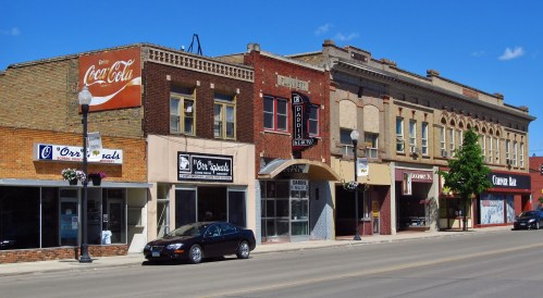 A small-town street featuring brick buildings, shops, and a classic theater under a clear blue sky.