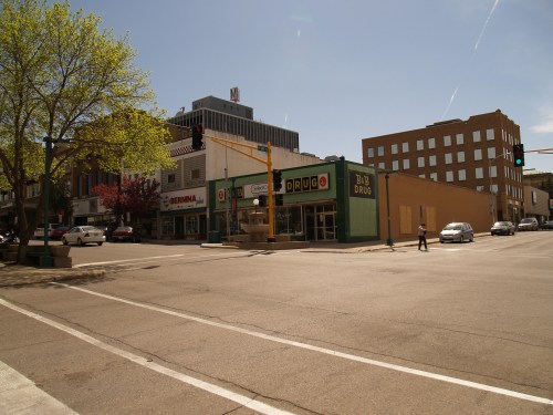 A street view showing shops, traffic lights, and pedestrians on a sunny day in a downtown area.