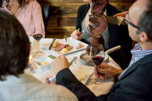 A group dining at a restaurant, enjoying a meat carving with wine and side dishes on the table.