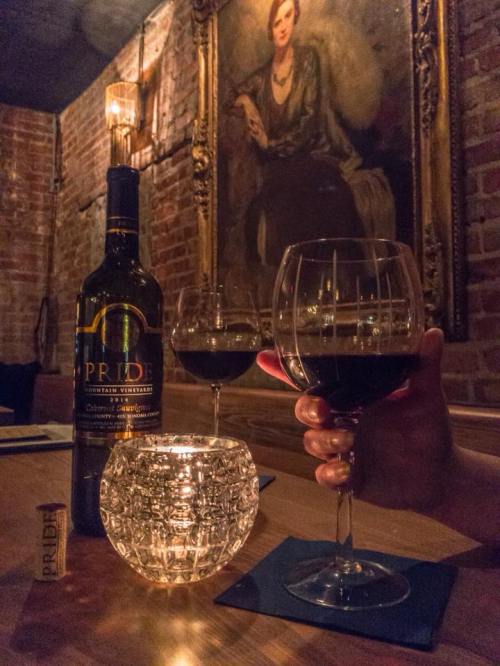 A dimly lit table with two glasses of red wine, a bottle of Pride wine, and a candle, set against a vintage portrait.
