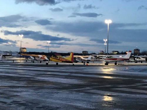 A row of parked airplanes at dusk, with city lights in the background and a cloudy sky.