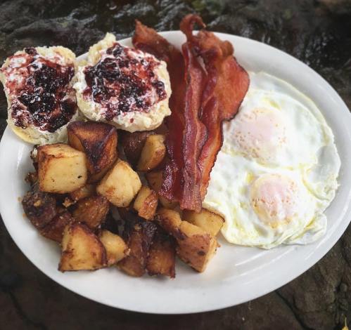 A plate of breakfast featuring two fried eggs, crispy bacon, roasted potatoes, and biscuits with jam.