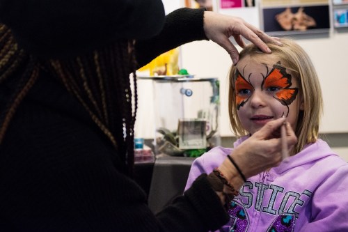 A child with butterfly face paint smiles as an artist adds details to her design.