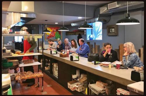 A busy café interior with patrons seated at a counter, enjoying food and drinks, while staff prepare meals in the background.