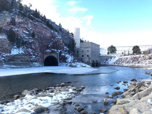 A snowy landscape featuring a river, rocky shore, and a large tunnel entrance beside a stone building.