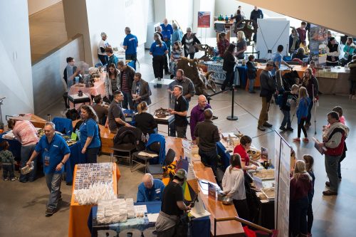 A busy event with people at tables, showcasing products and engaging in activities in a large indoor space.