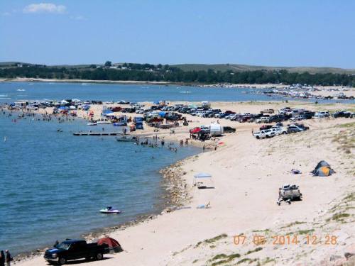 A busy beach scene with cars parked along the shore, people swimming, and tents set up on the sand.