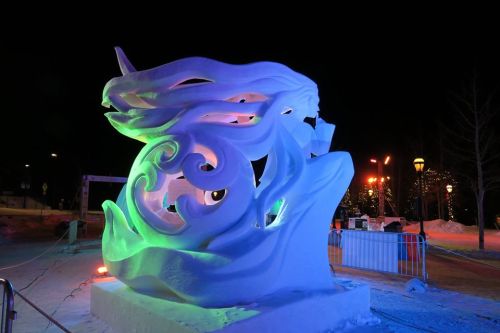 A large, illuminated ice sculpture with flowing designs, set against a night backdrop with colorful lights.