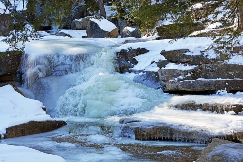A frozen waterfall cascades over rocky terrain, surrounded by snow-covered trees and icy formations.