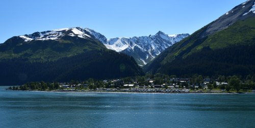 A serene landscape featuring snow-capped mountains, lush green hills, and a calm blue lake with a small town along the shore.