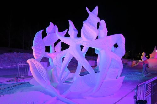 A colorful ice sculpture of abstract figures intertwined with large leaves, illuminated in purple and blue light at night.