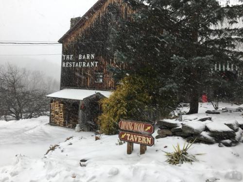 A rustic barn restaurant in a snowy landscape, with a sign for the dining room and tavern visible. Snow is falling.