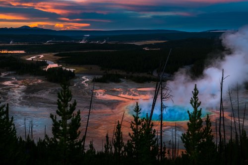 A vibrant sunset over Yellowstone, featuring steam rising from hot springs and a colorful landscape of trees and water.