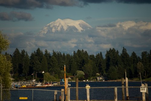 A scenic view of a lake with boats, surrounded by trees, and a snow-capped mountain in the background under a cloudy sky.