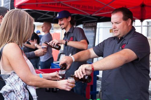 A vendor pours beer for a customer at a festival booth under a red tent, with others enjoying the event in the background.