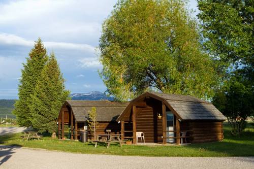 Two wooden cabins surrounded by trees, with a gravel path leading to them and mountains in the background.