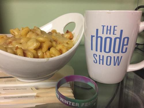A bowl of cheesy pasta next to a mug labeled "The Rhode Show" and a colorful wristband.