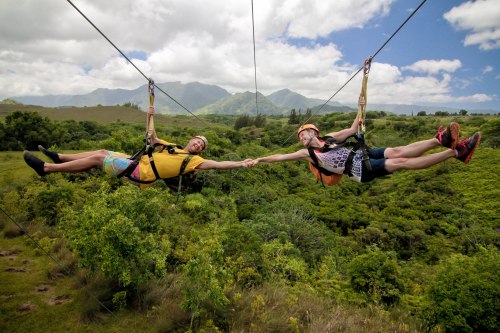 Two people ziplining over lush green hills, holding hands and smiling, with mountains in the background under a blue sky.