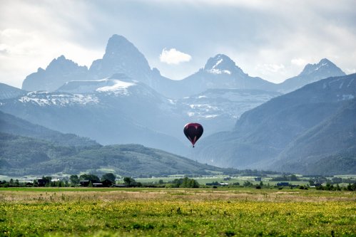 A hot air balloon floats over a lush valley with snow-capped mountains in the background under a cloudy sky.