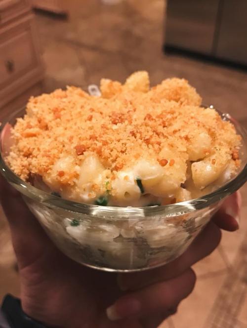 A hand holding a glass bowl of macaroni and cheese topped with golden breadcrumbs.
