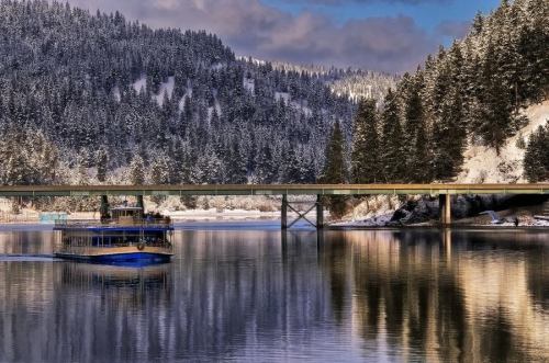 A boat glides on a snowy river, with a bridge and evergreen trees reflected in the calm water.