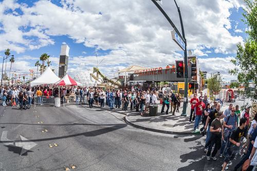 A large crowd gathers outside a venue with tents and palm trees under a partly cloudy sky.