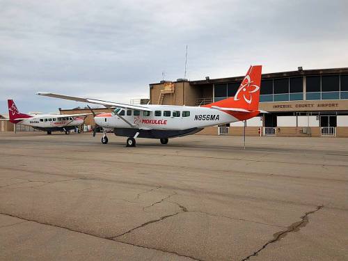 A small airplane with orange and white markings parked at Imperial County Airport, with another plane in the background.