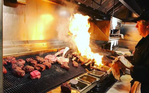 A chef grilling various cuts of meat over an open flame in a busy kitchen. Flames rise dramatically from the grill.