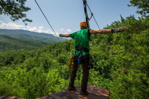 A person in a green shirt and helmet stands on a platform, arms outstretched, overlooking a lush mountain landscape.