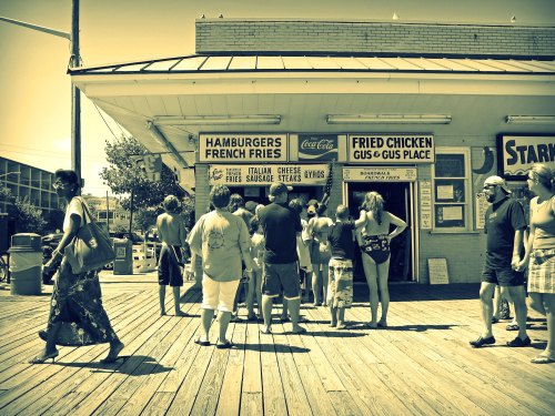 A busy outdoor food stand with people in line, surrounded by a sunny boardwalk atmosphere.