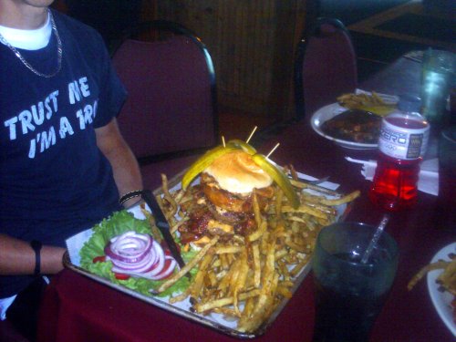 A large burger topped with pickles and onion, served on a bed of fries, with a person wearing a "Trust Me, I'm a Pro" shirt.