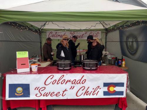 A tent with a "Colorado Sweet Chili" sign, featuring people serving chili and decorations, including Colorado flags.