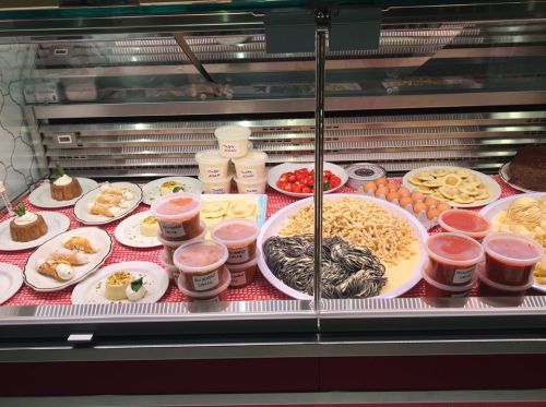 A display case filled with various dishes, including pasta, desserts, sauces, and fresh ingredients.