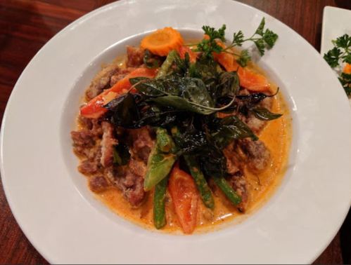 A plate of curry with crispy meat, green beans, carrots, and herbs, garnished with fresh greens on a wooden table.